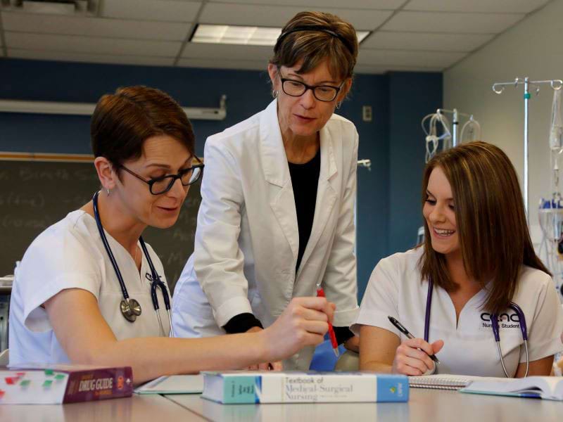 An instructor guides CCAC Nursing students wearing scrubs as they pore over papers on a desk.