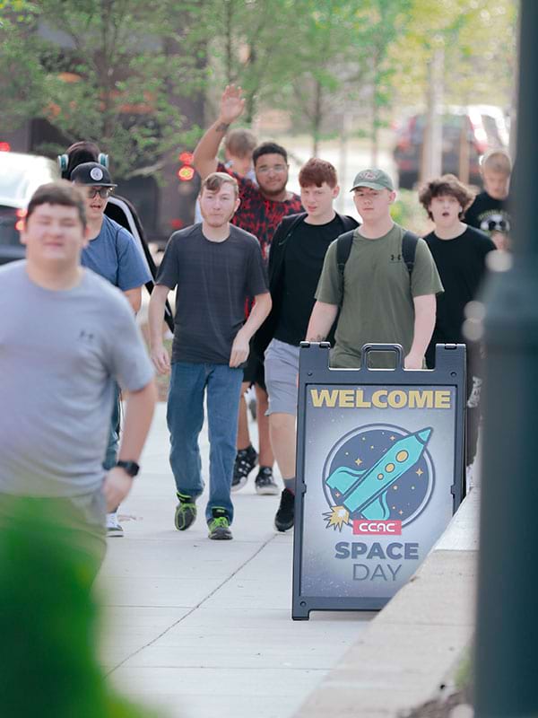 Students arriving for Space Day with a sidewalk sign saying Welcome CCAC Space Day with an illustration of a rocket ship