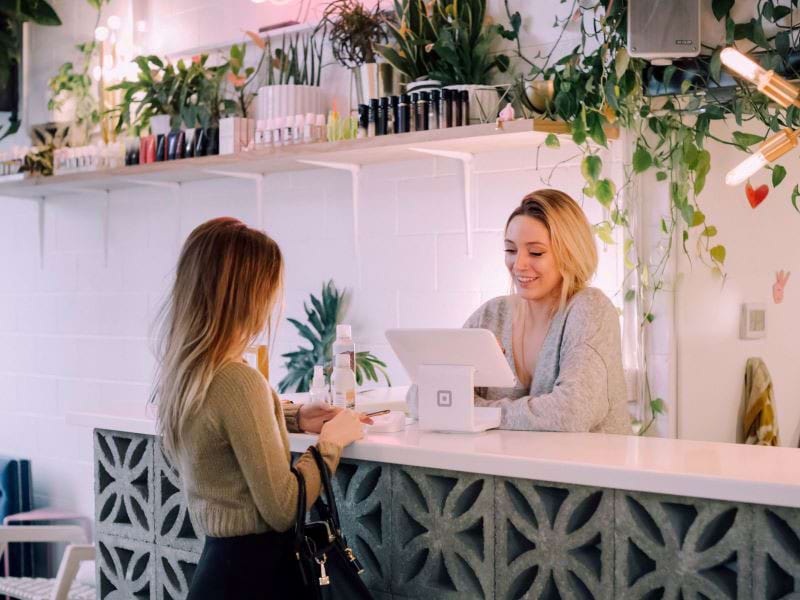 A shopkeeper smiles as she helps a customer pay for a purchase in a small shop.